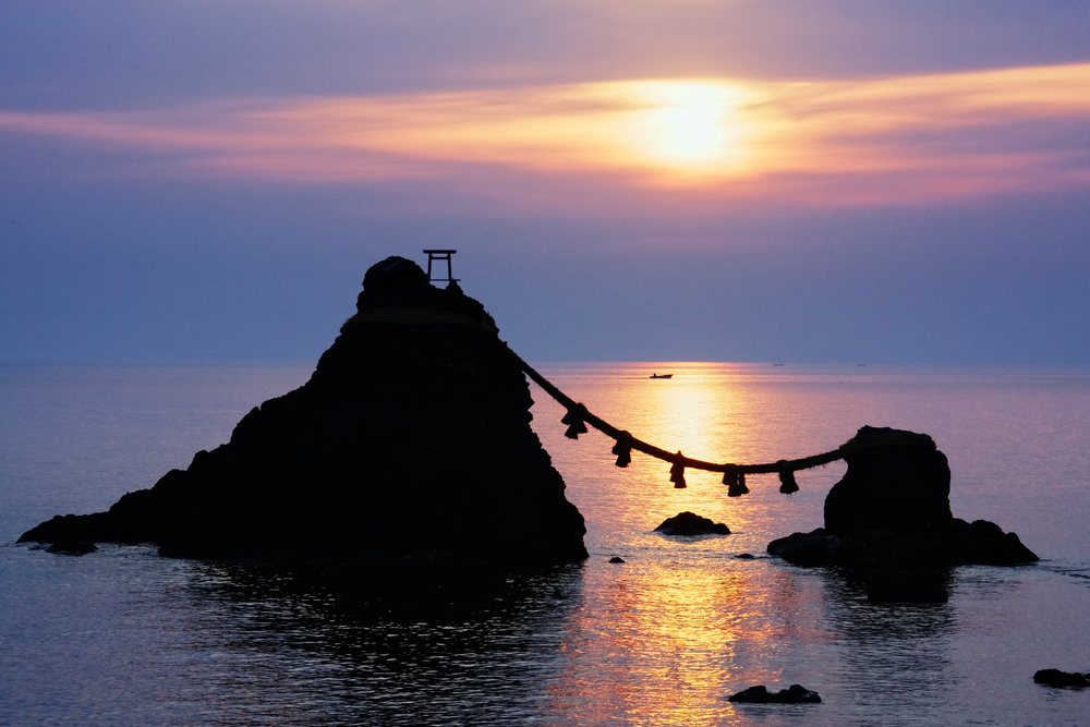 Sunset over Meoto Iwa rock formation and torii gate silhouette on calm ocean waters, Honshu, Japan.