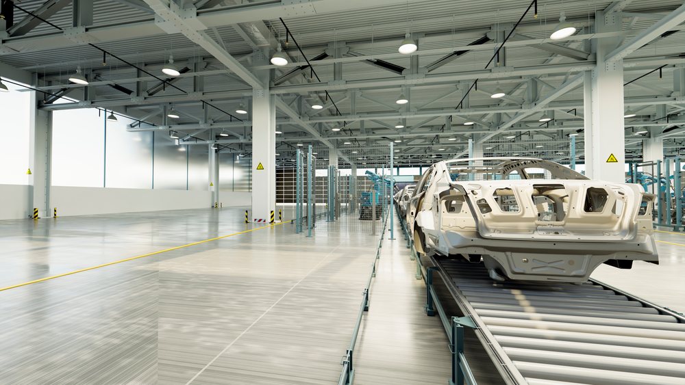 Car frames move along an assembly line inside a clean, spacious automotive manufacturing plant with robotic arms and industrial lighting.
