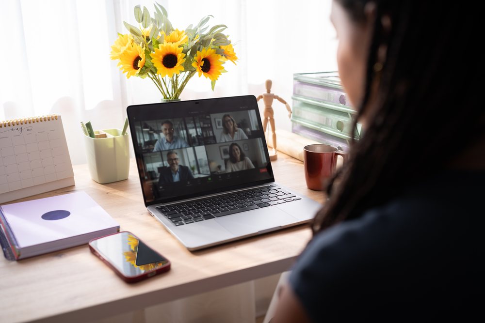 A woman participates in a video conference on a laptop in a sunlit home office, with a desk decorated with sunflowers and office supplies, remote work, student, Spain.