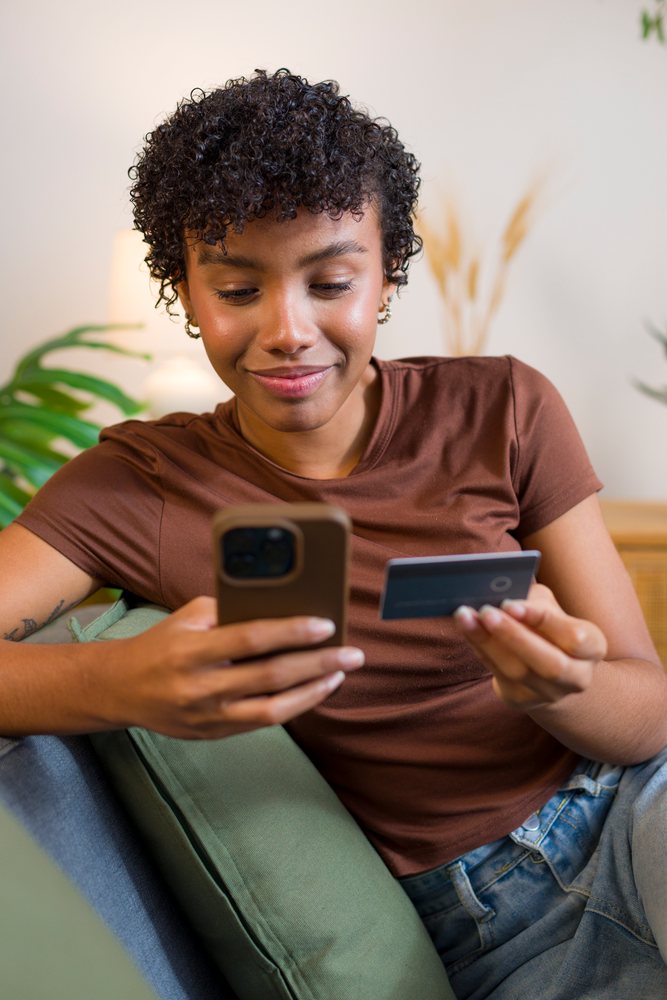 Young woman with curly hair sitting on a couch, smiling while holding a smartphone and credit card, possibly shopping online.