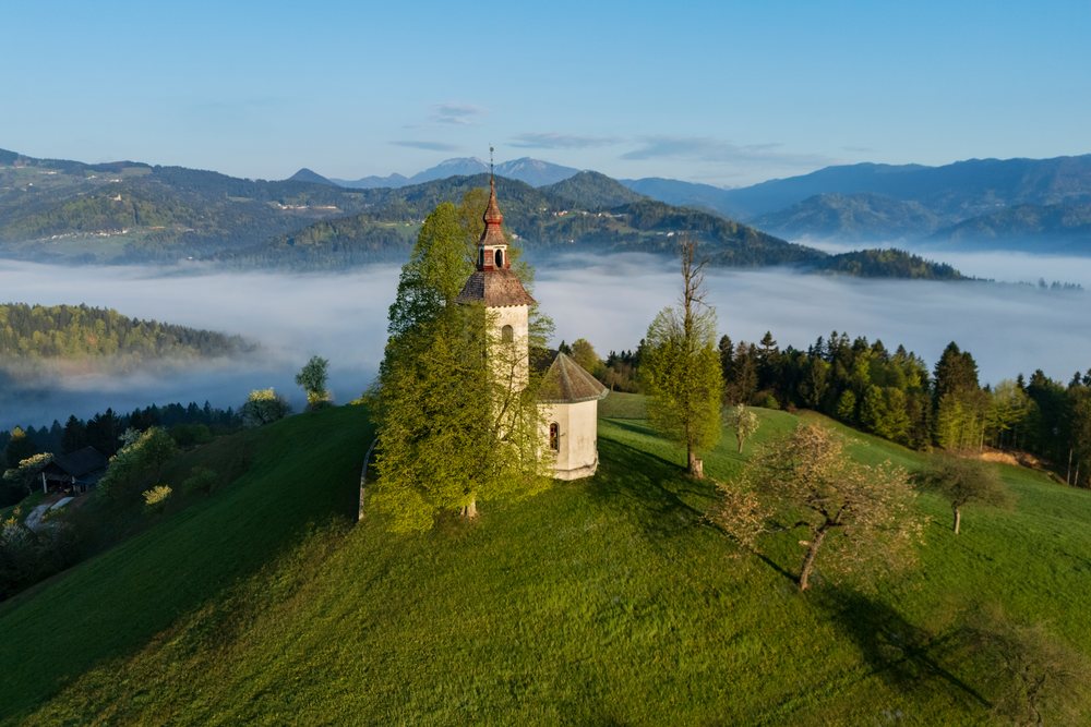 Church on a green hilltop surrounded by trees with fog in the valley and mountains at Sunrise, landscape, cityscape, Skofje Loka, Slovenia.