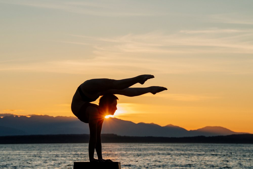 Woman performing a balanced handstand on a pedestal by water and mountains at sunset, USA.