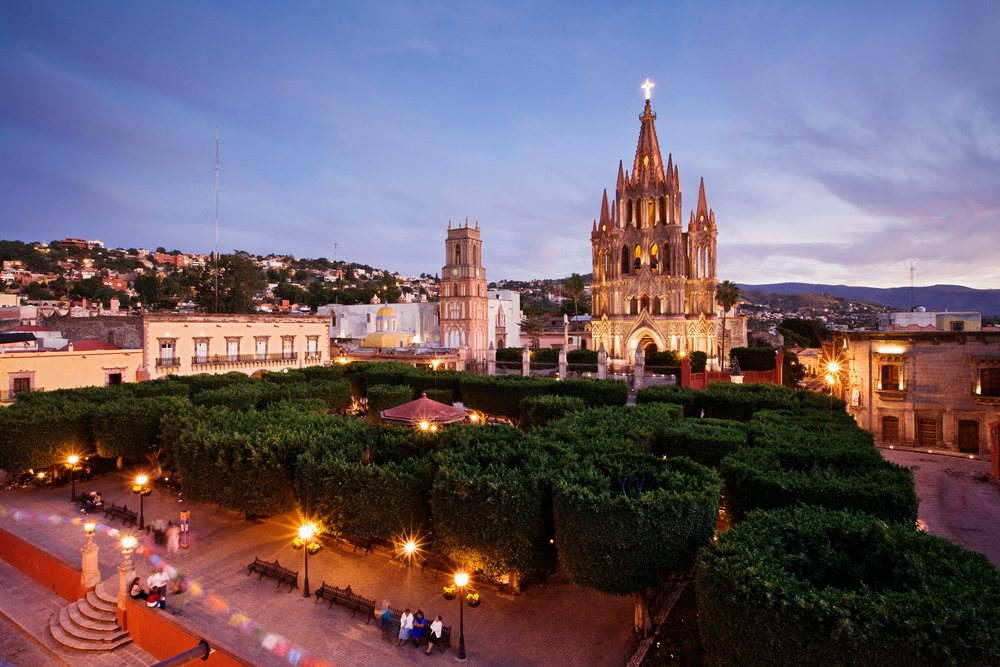 San Miguel de Allende shows an illuminated church with spires beside a manicured park with trees and benches, Dusk, Mexico.