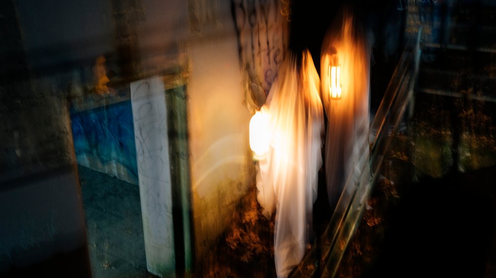 Two people dressed as ghosts with one holding a lantern stand in a dark hallway near metal stairs and a wall with blurred motion effects, USA.
