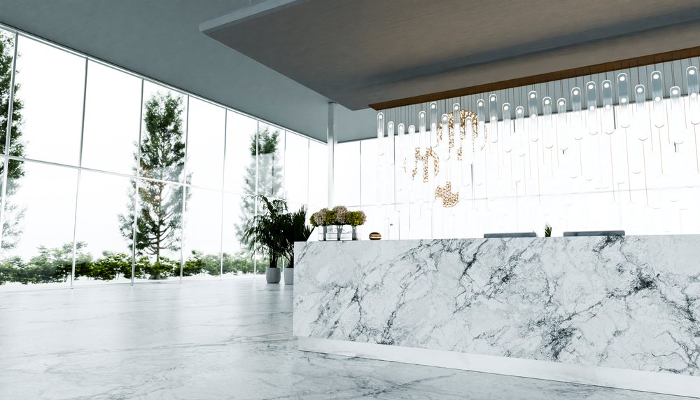 Spacious modern lobby featuring a marble reception desk with decorative flowers, indoor plants, and a contemporary chandelier under natural daylight.