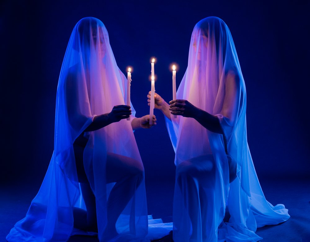 Two adult figures kneeling and each covered with a sheer veil hold lit candles in a blue-lit dark studio, ghosts, USA.