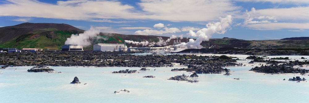 Steam rises from a geothermal power plant amid volcanic rocks and blue geothermal water's volcanic landscape under a partly cloudy sky.