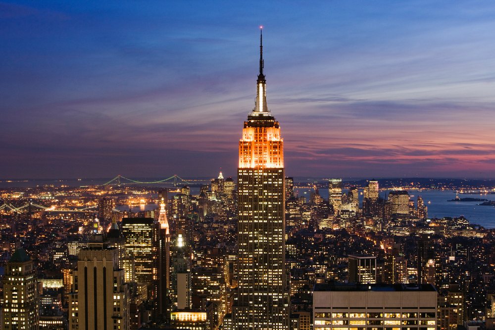 Empire State Building lit up at dusk with New York City skyline and bridges visible in the background over water, USA.