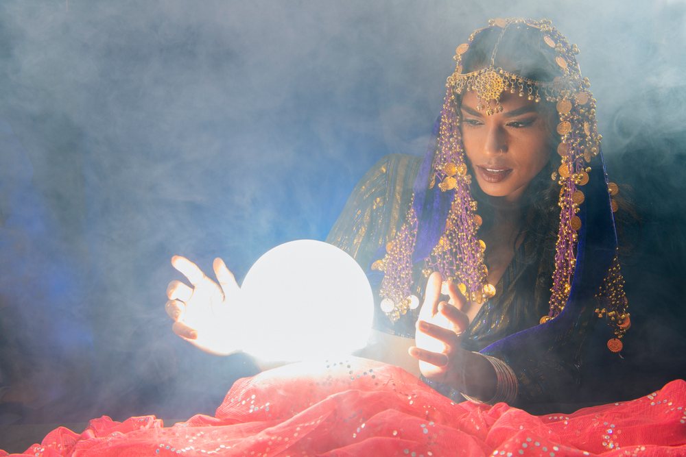 A fortune teller dressed in a purple veil with gold coins gazes intently at a glowing crystal ball surrounded by red fabric and smoke in a mystical indoor setting in the, USA.