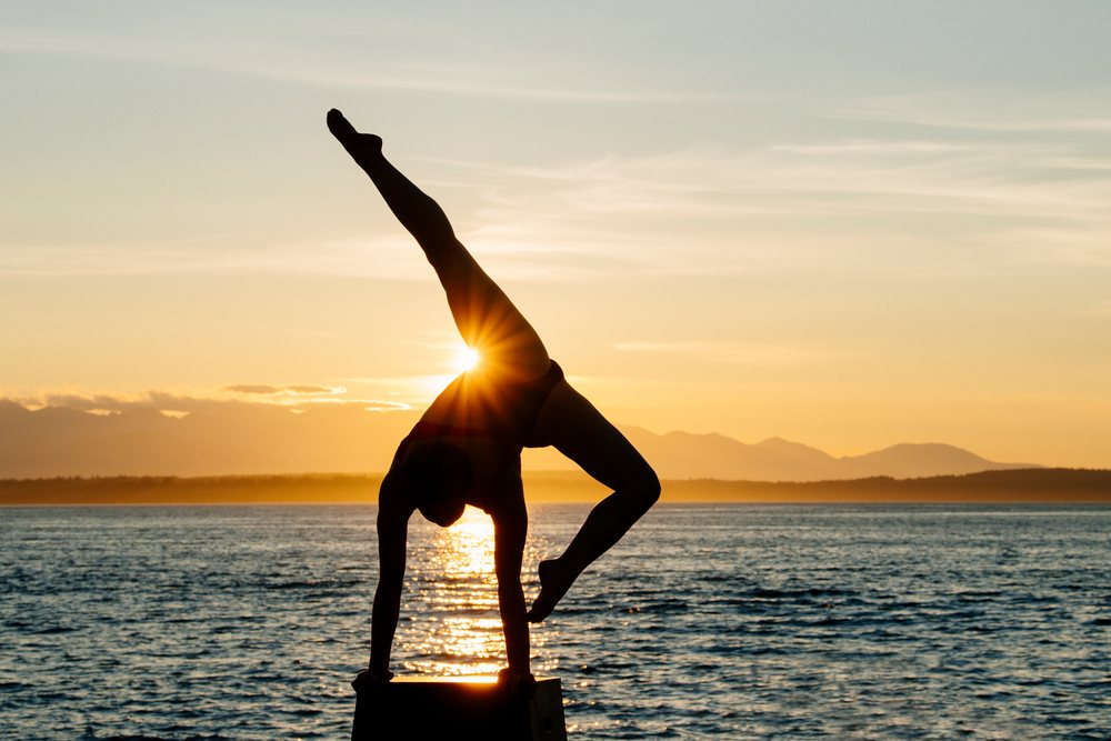 Silhouette of a woman performing a handstand on a platform by the ocean at sunset with mountains in the background, USA.