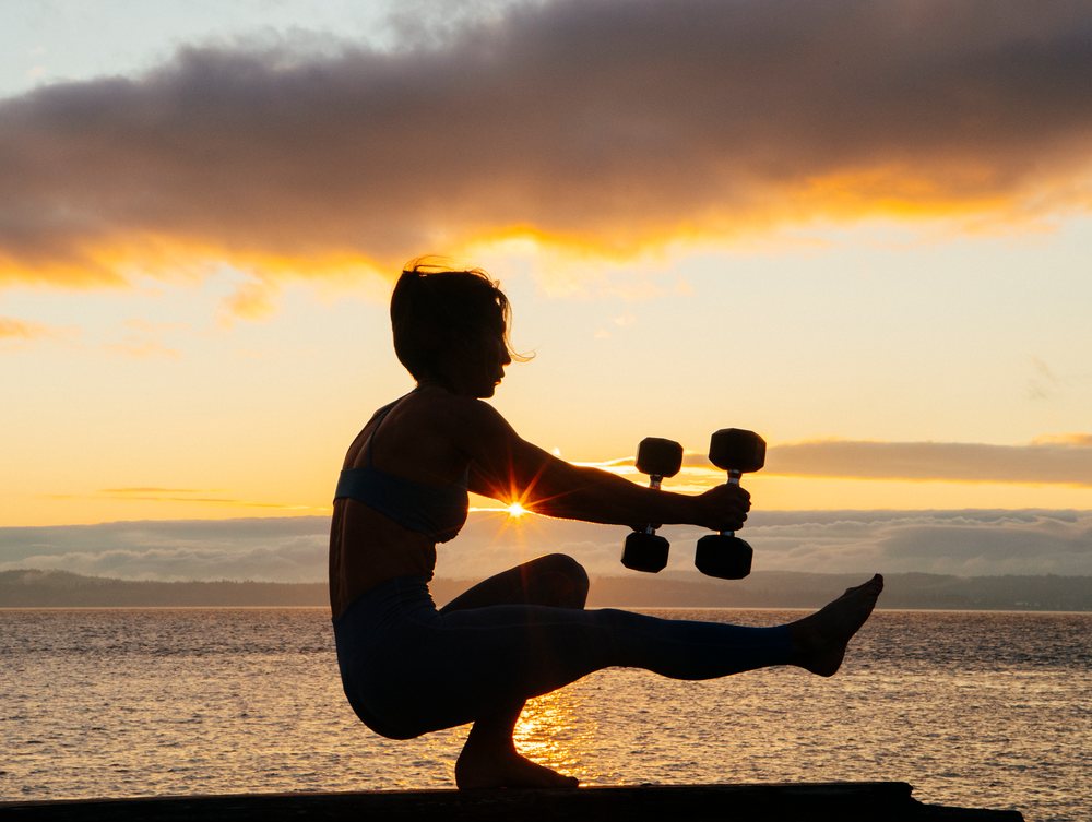 Woman performing balancing yoga pose with dumbbells outdoors at sunset by a water body, WA, USA.