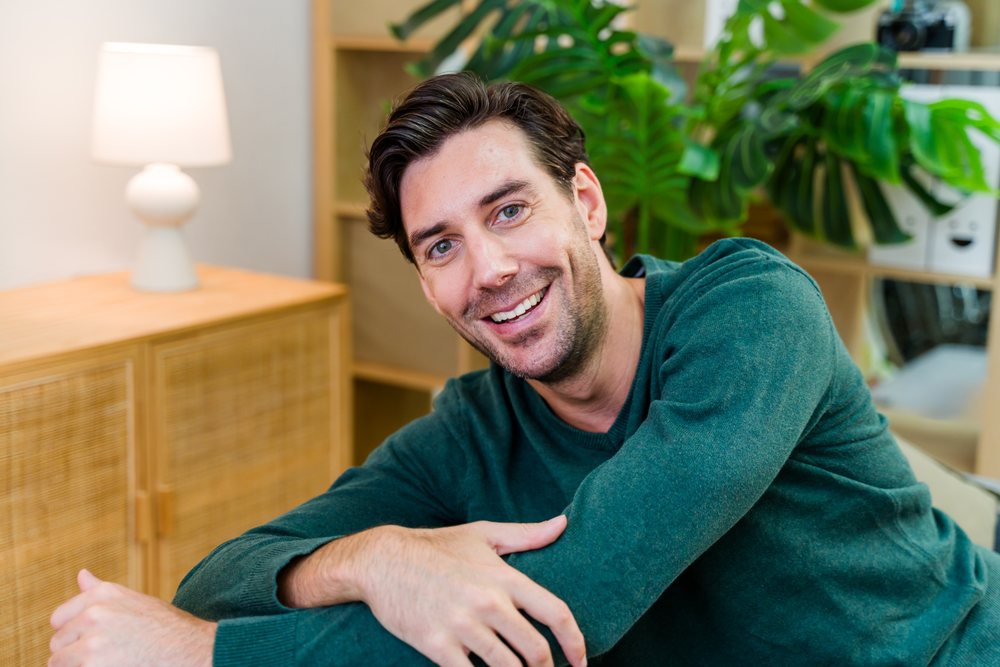 Man wearing a green sweater is sitting and smiling indoors in a home living room with plants and a lamp on a cabinet, Spain.