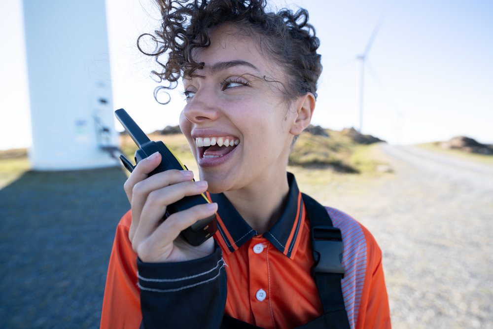Smiling woman wearing an orange work shirt and black harness talks on a walkie-talkie beside a wind turbine at a wind farm during daytime, sustainability, renewable energy, Galicia, Spain.