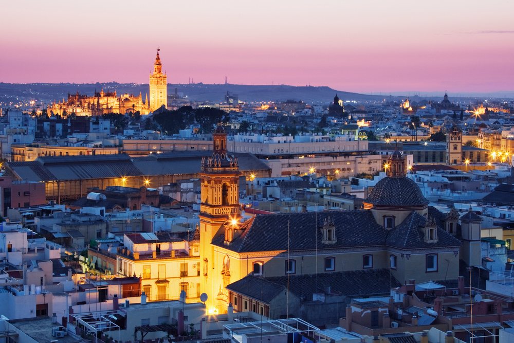 Cityscape of Seville showing illuminated Giralda bell tower and cathedral under a purple and pink sky, Andalucia, Spain.