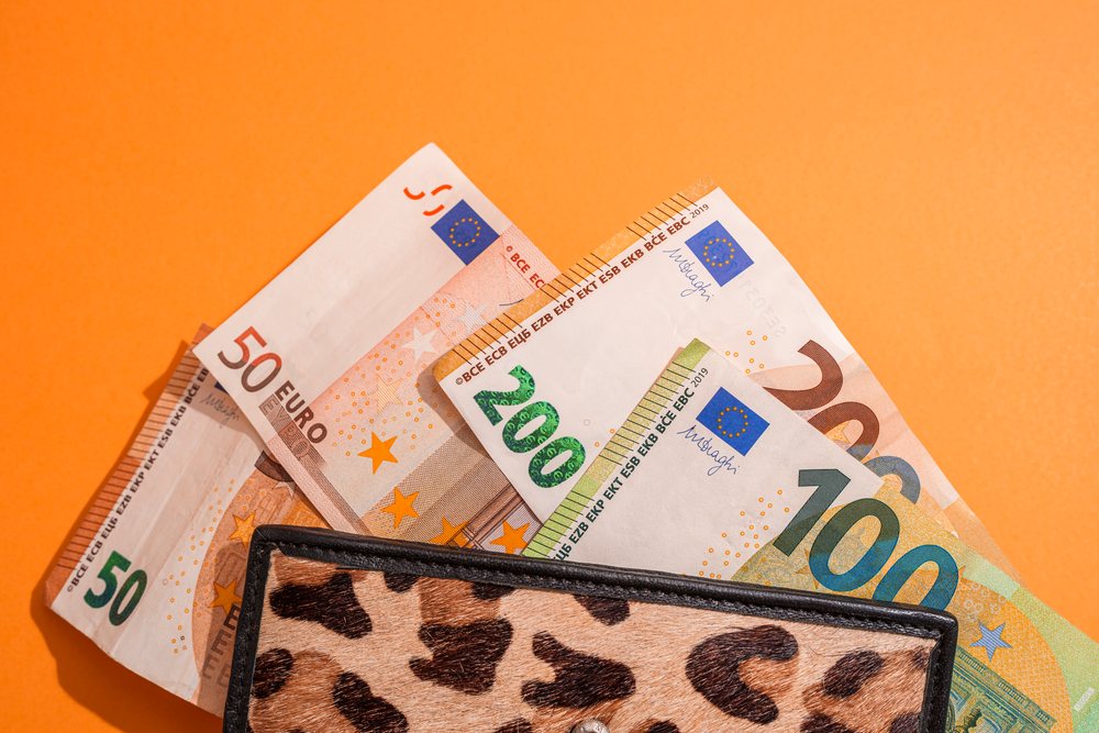 Euro banknotes in denominations of 20, 50, 100, and 200 partially protruding from a leopard print wallet against an orange studio background, Studio Shot.