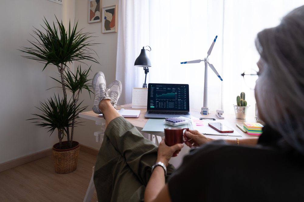 Person relaxing at a home office desk in Spain holding a coffee cup with a laptop displaying stock market charts.