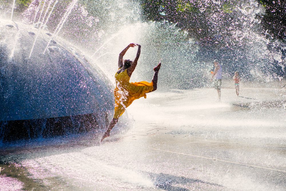 Woman in a yellow dress jumps and splashes water near a large outdoor fountain on a sunny day in a public urban park, USA.