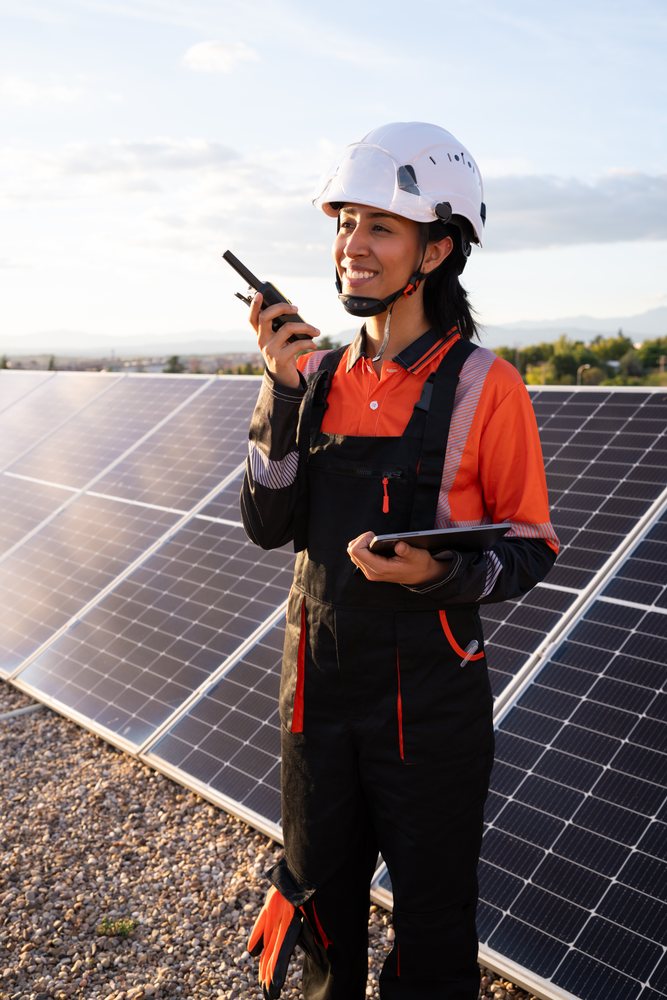 Female technician wearing safety gear uses a walkie talkie and tablet while inspecting solar panels outdoors, Madrid, Spain.