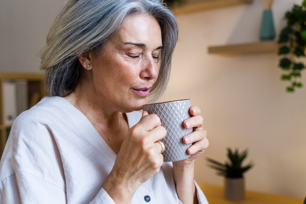 Mature woman with grey hair holds a grey textured ceramic mug and drinks a warm beverage indoors with a peaceful expression, Spain.
