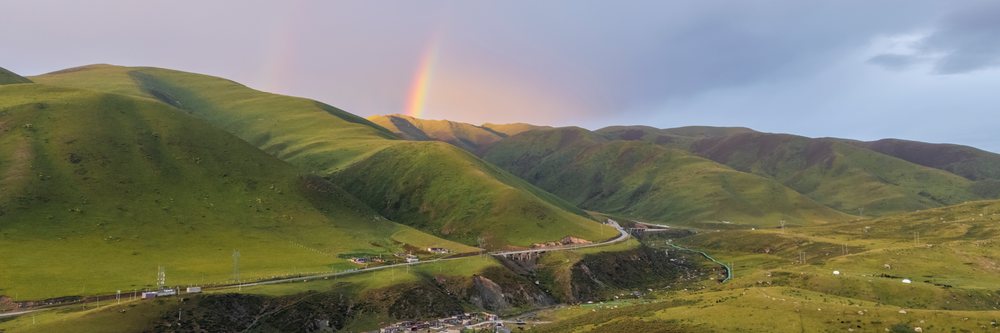 Sunset view of green mountainous landscape with rainbow over hills and a village near Dege Zhuqing Mansion Hotel in Kham Tibet, China, Zhuqingcun, Sechuan Province, People's Republic of China.