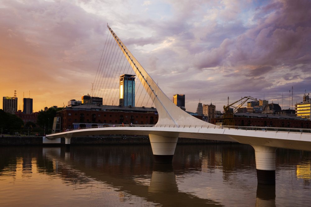 Puente de la Mujer, a modern cable-stayed pedestrian bridge over a river with urban buildings reflecting in the water at sunset, Buenos Aires, Argentina.