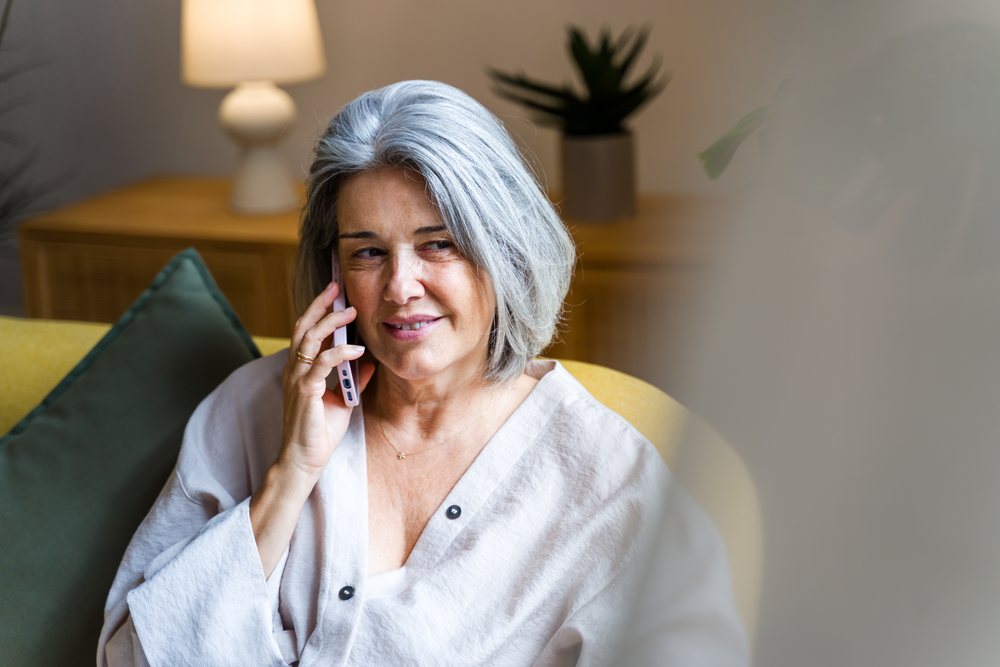Middle-aged woman with gray hair talking on the phone while sitting on a sofa in a cozy home interior with lamp and indoor plant, Spain.