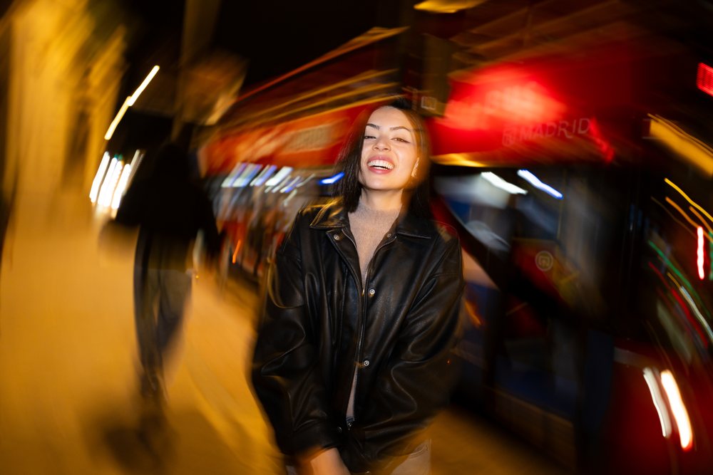 Smiling woman with dark hair wears a leather jacket standing on a nighttime urban street with people walking and a red bus behind her, Madrid, Spain.
