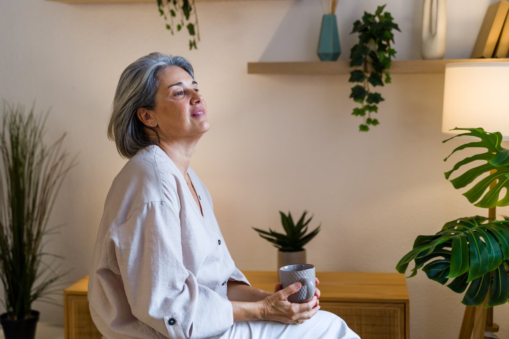 Middle-aged woman with gray hair sitting indoors holding a mug in a peaceful home interior with plants and a table lamp, Spain.