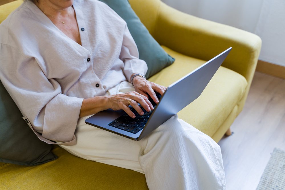 Woman typing on a laptop while seated on a yellow couch in a living room with wooden flooring, Spain.