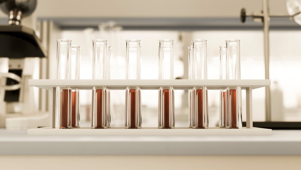 Test tubes filled with brown liquid arranged in a rack on a laboratory bench with scientific equipment visible in the background.