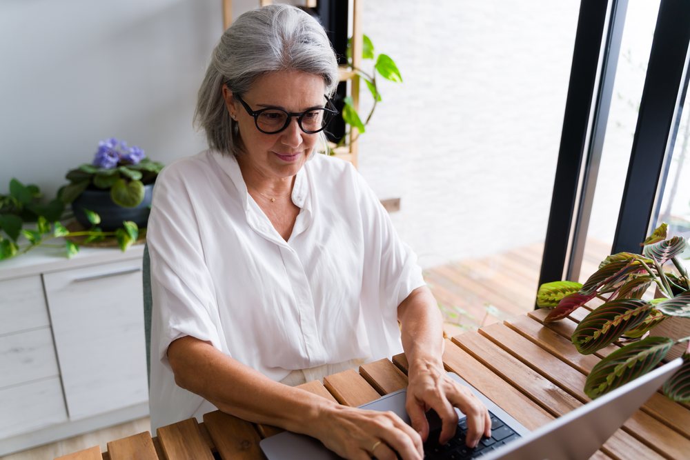Woman with grey hair typing on a laptop at a wooden desk with indoor plants in a bright home office, Spain.