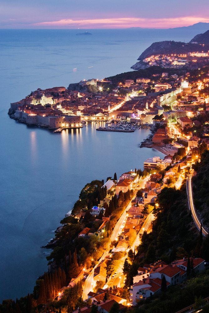 Historic Old City of Dubrovnik illuminated, showing sea coastline, harbor with boats, and stone city walls under a colorful sky, Dusk, Croatia.