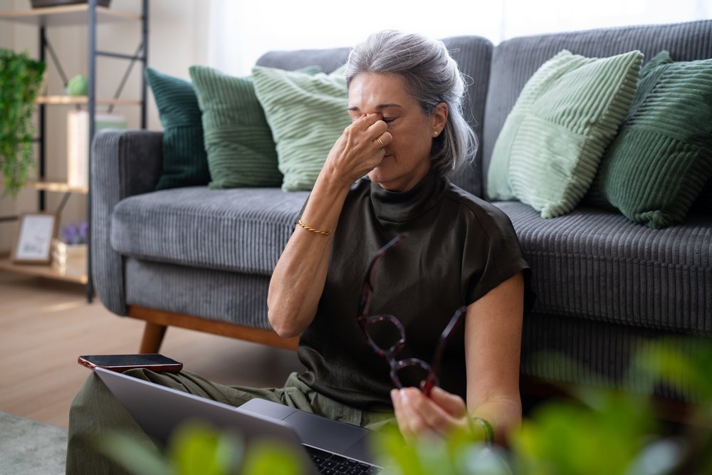 Woman with gray hair seated on a living room floor rubbing her eyes near a couch with green pillows and holding eyeglasses, Spain.