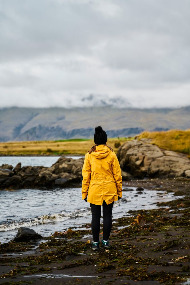 A white woman stands on a rocky shore with seaweed