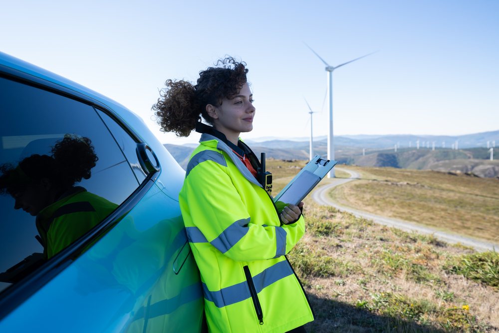 Woman engineer in hi-vis jacket with clipboard inspects wind turbines on a renewable energy wind farm, Galicia, Spain.
