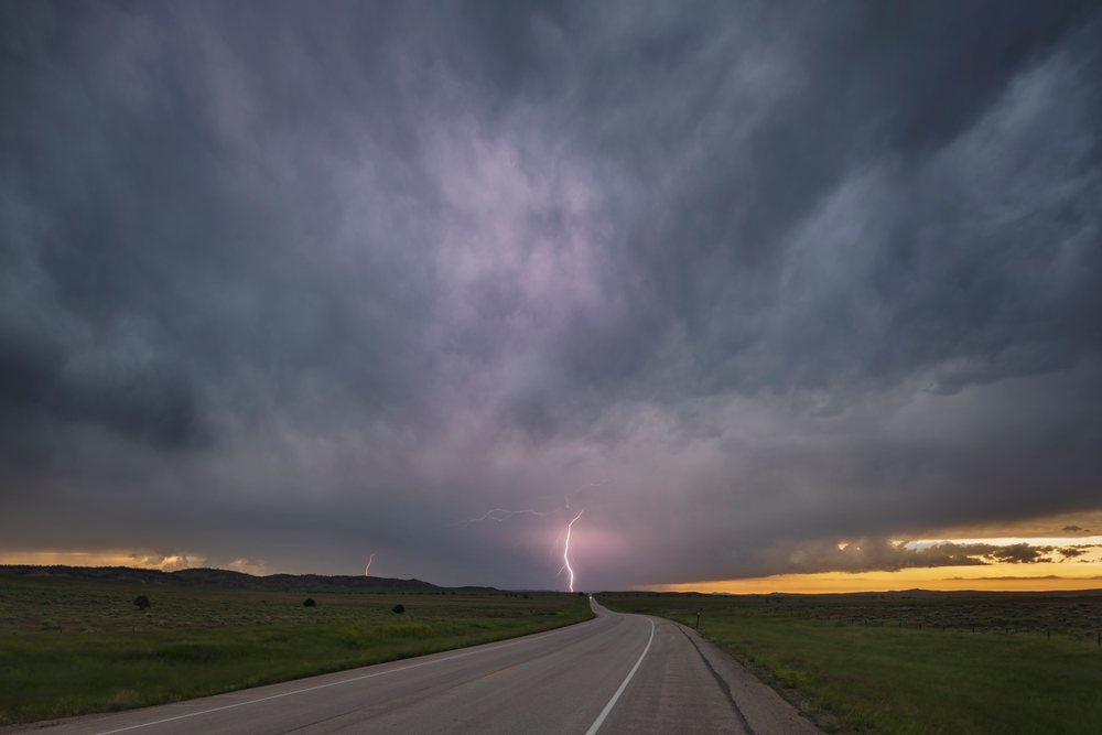 Lightning strikes near the horizon over a curved Highway across grassland fields under storm clouds and a Sunset sky in Wyoming's Great Plains, Gillette, USA.