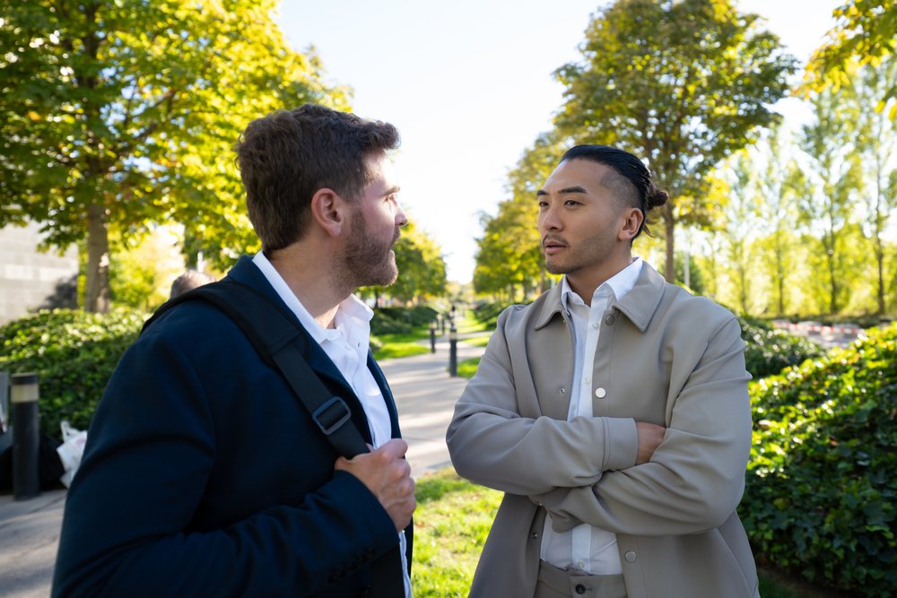 Two men wearing casual business attire are conversing while standing on a sunny park pathway, Madrid, Spain.