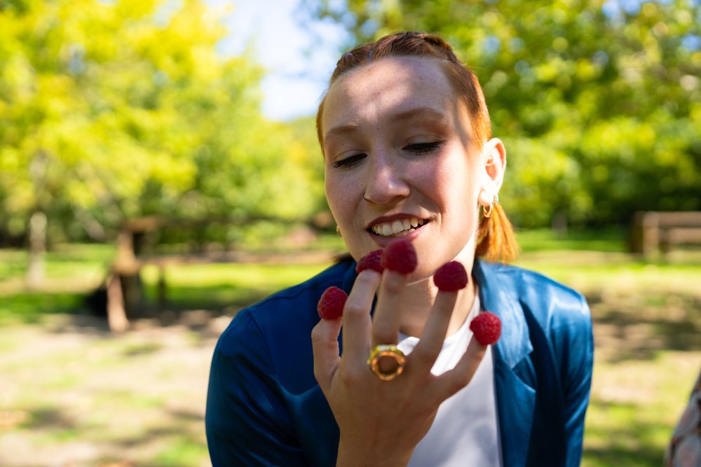 Woman smiling outdoors in a park while holding raspberries on her fingers with a green tree background, Spain.