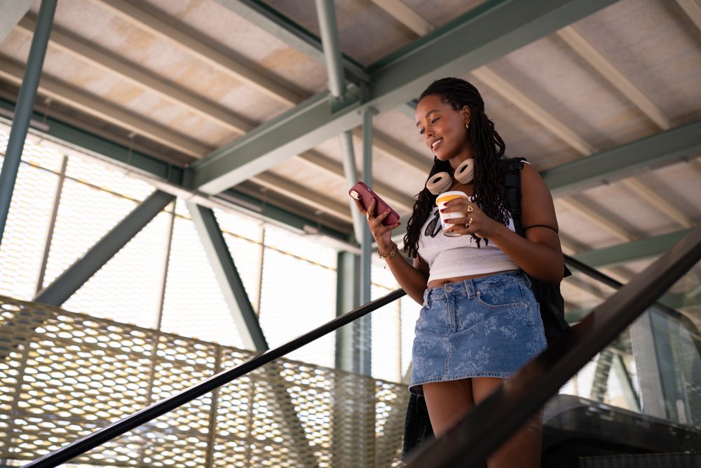 A young woman standing on an escalator uses her smartphone and holds a coffee cup inside a modern building, remote work, travel, Santiago De Compostela, Spain.