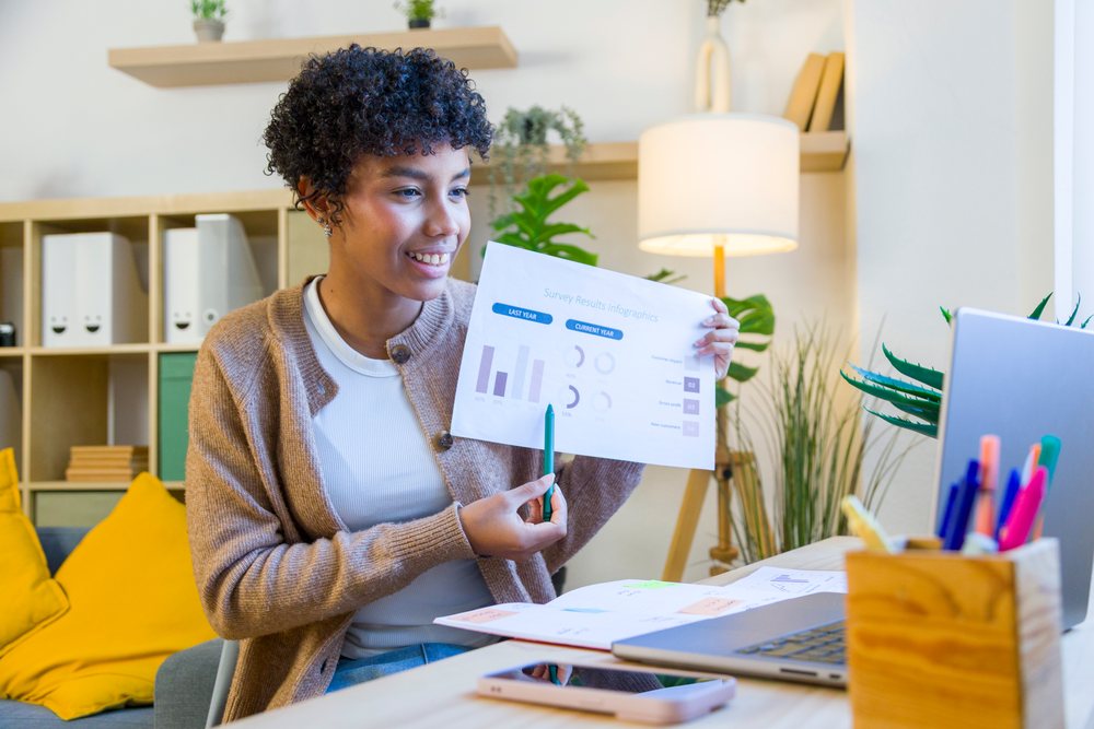 Woman sitting at a home office desk holding a pen and paper with survey result infographics, with laptop and smartphone on the desk, remote work.