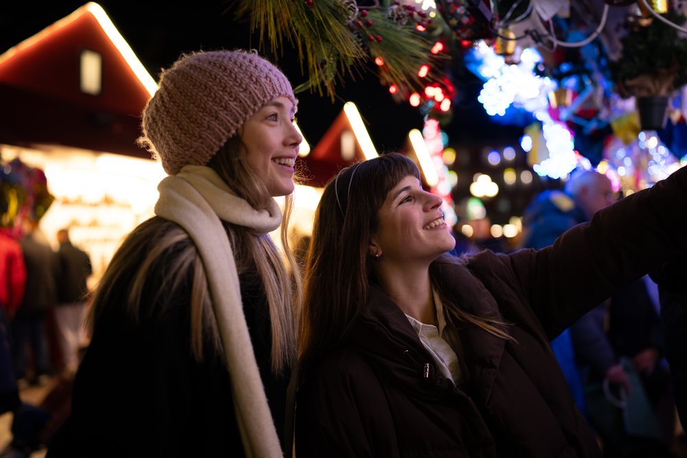 Two women smiling outdoors at a festive holiday market during the Christmas season at night, Madrid, Spain.