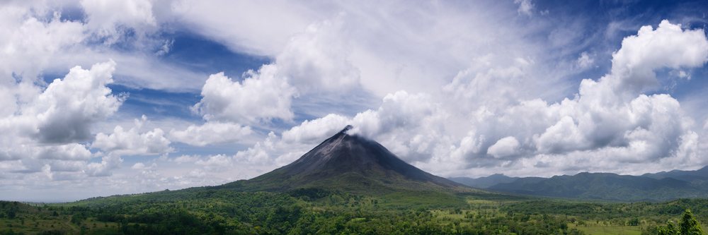 Arenal Volcano rises above green forested land under a partly cloudy sky.