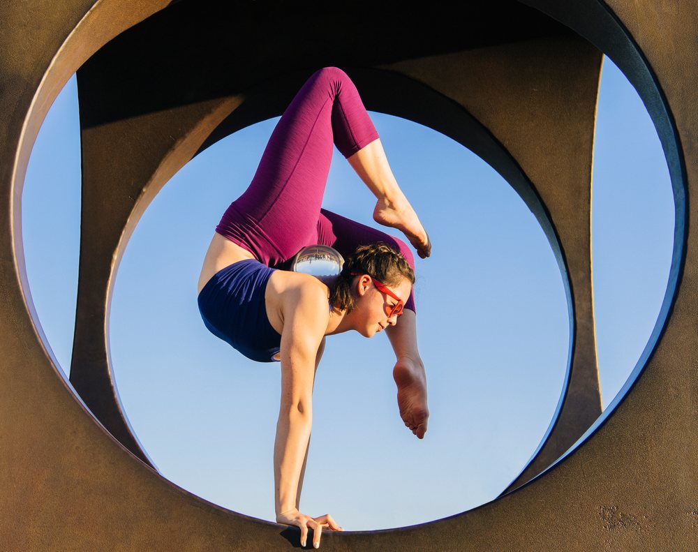 Woman in navy tank top and purple leggings performs a yoga handstand balanced inside a circular outdoor sculpture against a clear blue sky, USA.