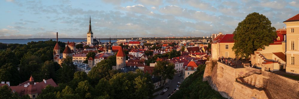 City skyline of Tallinn at sunset showing historic buildings, fortress walls with battlements, church steeple, and water beyond under a cloudy blue sky, cityscape, Estonia, Baltic States.