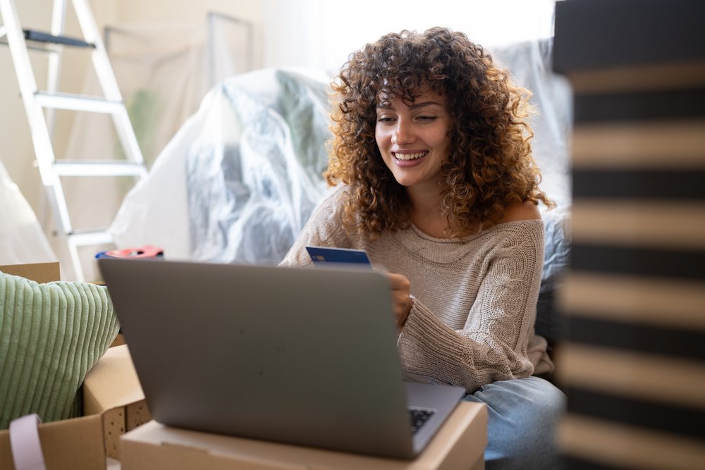 Woman with curly hair sits indoors among moving boxes using a laptop and a credit card, Spain.