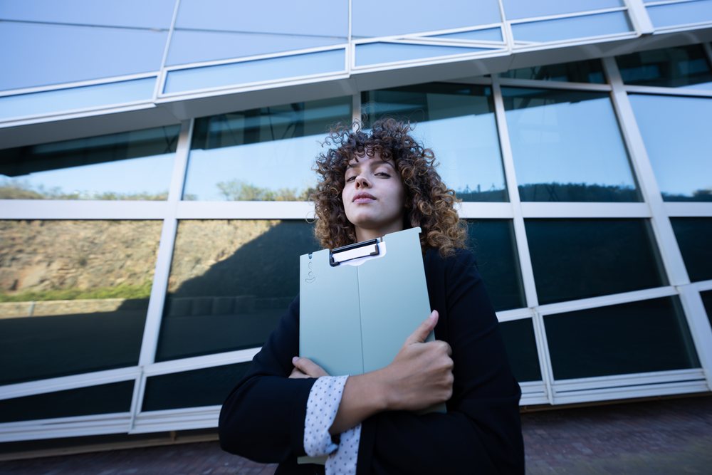 Businesswoman holding a clipboard standing in front of a modern office building with glass windows, business trip, Spain, Santiago De Compostela.
