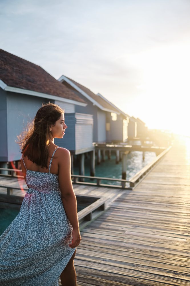 A woman in a summer dress walks along a wooden boardwalk past overwater bungalows at sunset in the, Maldives.