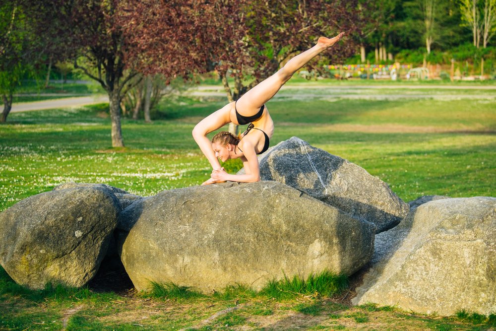 Woman in a black bikini performs a flexible yoga pose on a large rock in a sunlit green park, USA.