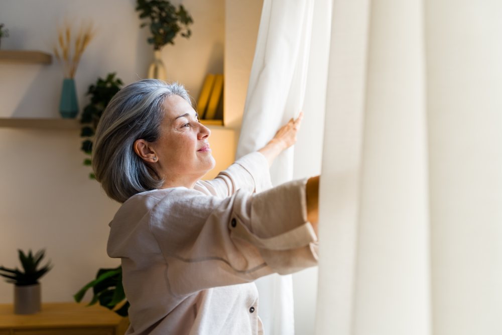 Middle-aged woman with gray hair opens curtains to welcome natural light into a cozy home interior with plants and decor, Spain.
