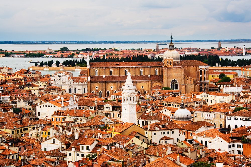 Overview of Venice cityscape showing orange and red rooftops, historic churches with dome and tower, and canals under a cloudy sky.